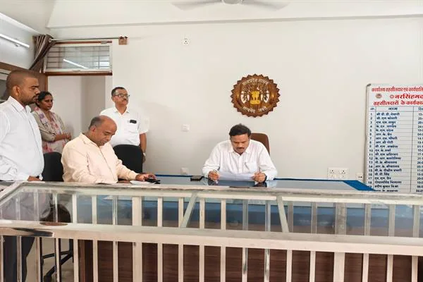 कार्यालय में मेज पर दस्तावेज देखते दो अधिकारी, पास खड़े कर्मचारी और सामने धातु की रेलिंग
